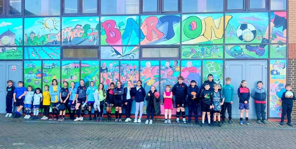 Barton United Football Team of children stood in front of colourful painted wall.